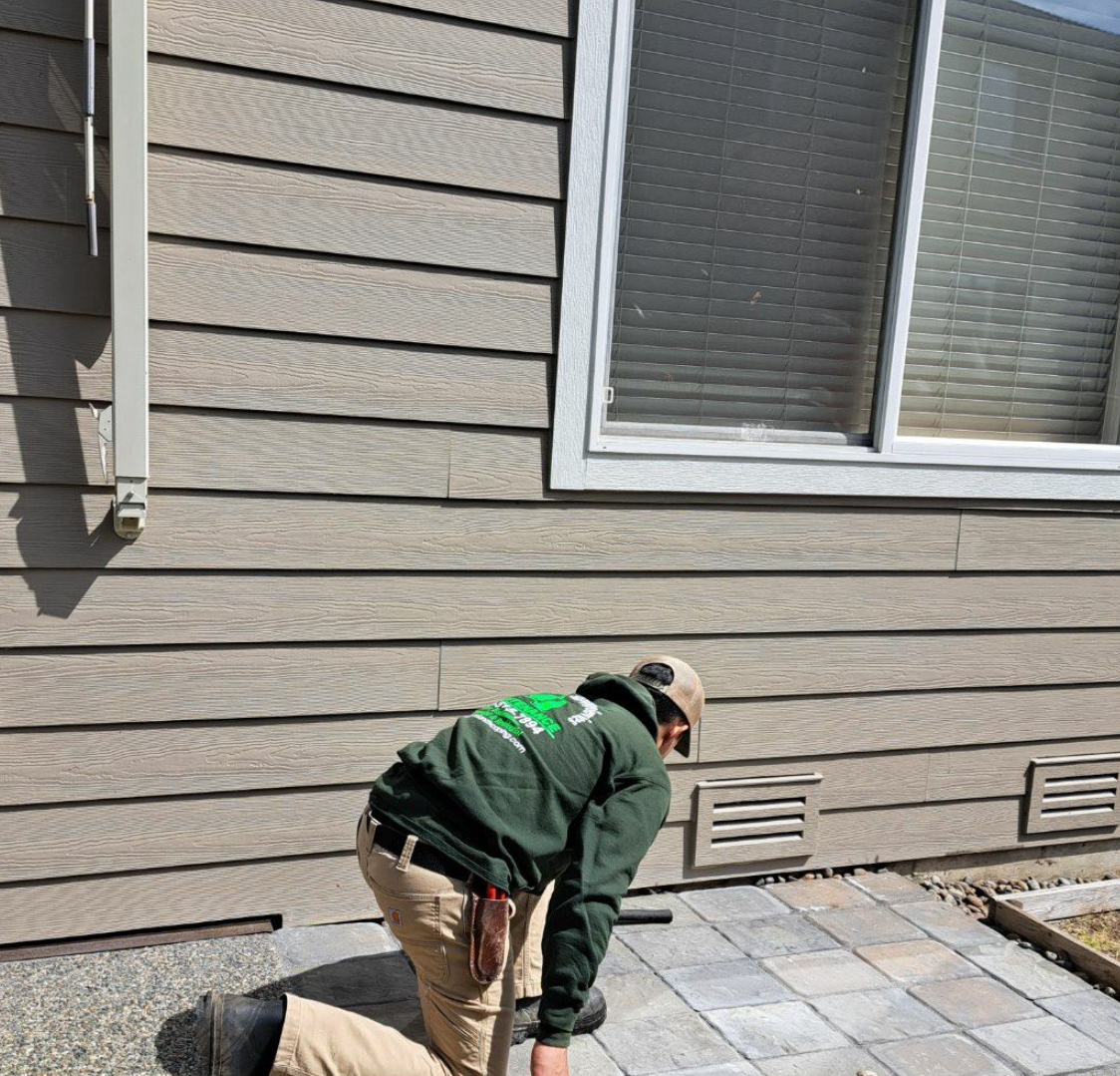 Landscaper installing stone pavers beside a house with beige siding.