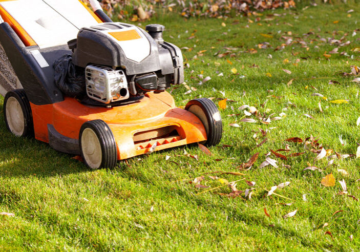 Close-up of an orange lawn mower cutting grass with scattered leaves.