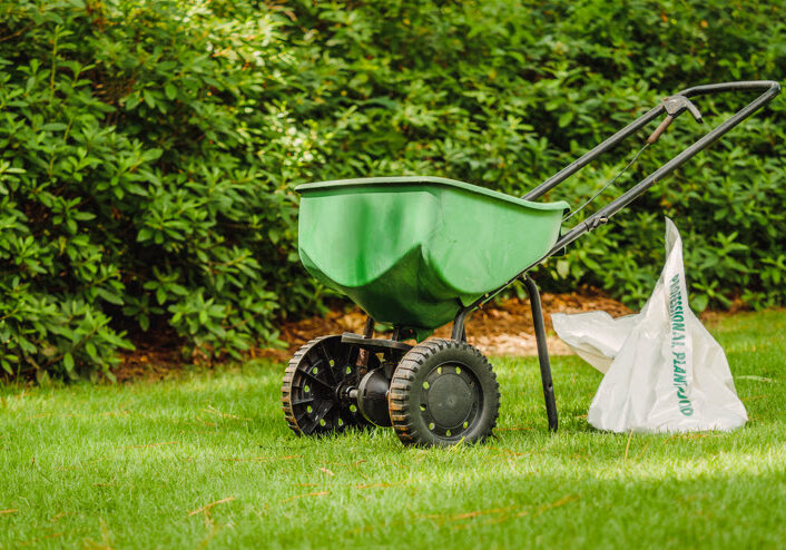 A green broadcast spreader sits on a well-manicured lawn next to a partially opened white bag.